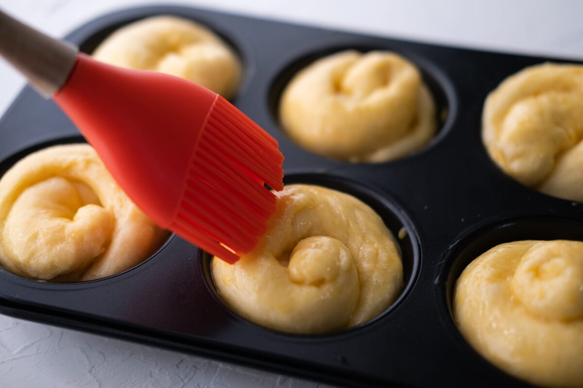 Brioche buns in a muffin pan brushed with egg wash.