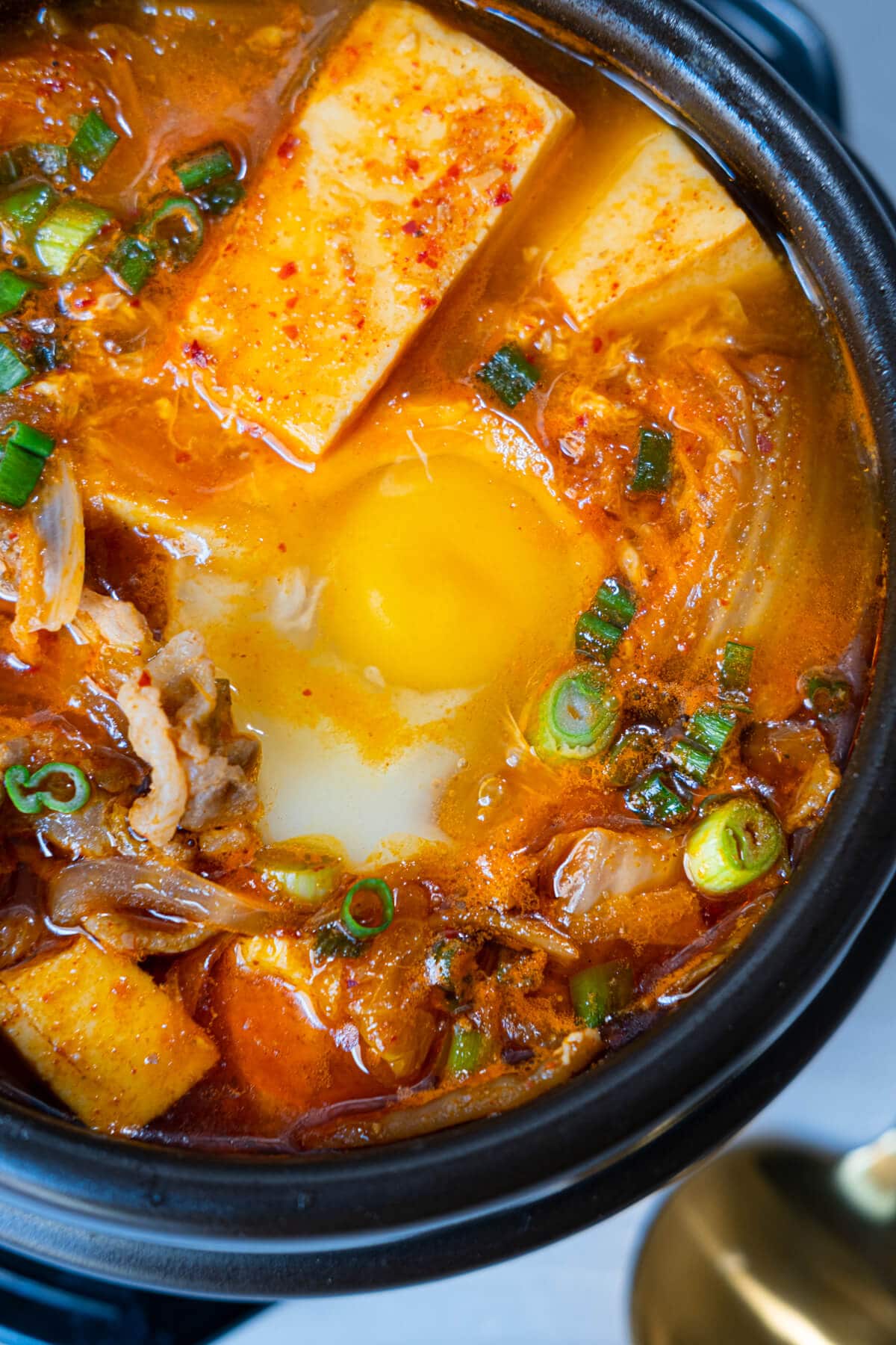 A close up shot of authentic Korean tofu stew made with pork belly, kimchi, and soft tofu served in a black clay pot. 