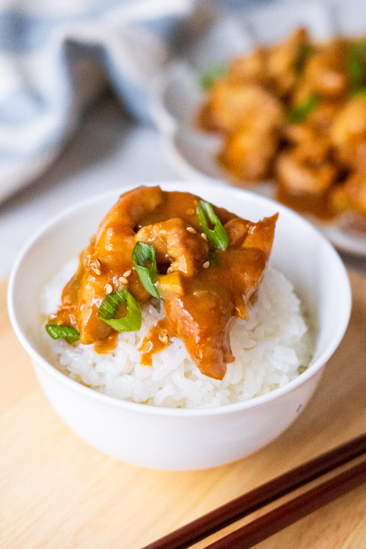 A bowl of rice served with chicken coated with orange bourbon sauce on top of it in a white bowl with chopsticks on the side. 