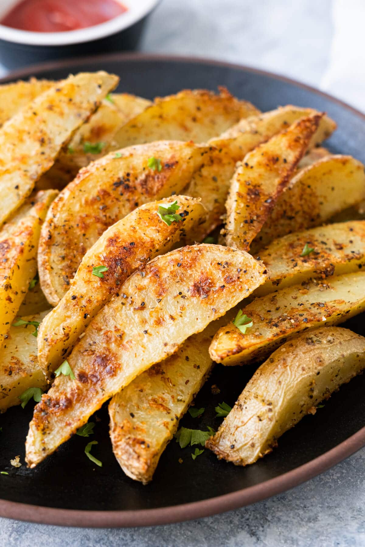 Potato wedges seasoned with garlic and parmesan cheese on a black plate with ketchup sauce served next to it. 