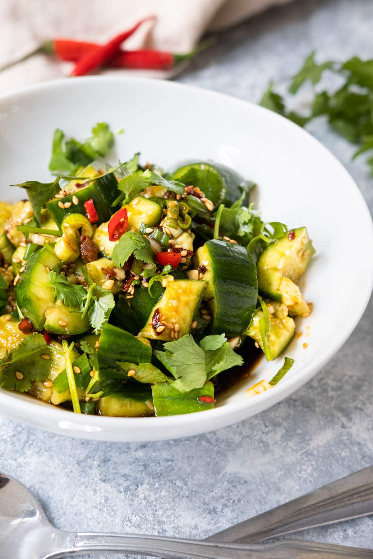 A small bowl of spicy cucumber served with tingly spicy salad dressing with 2 red pepper and cilantro placed behind.