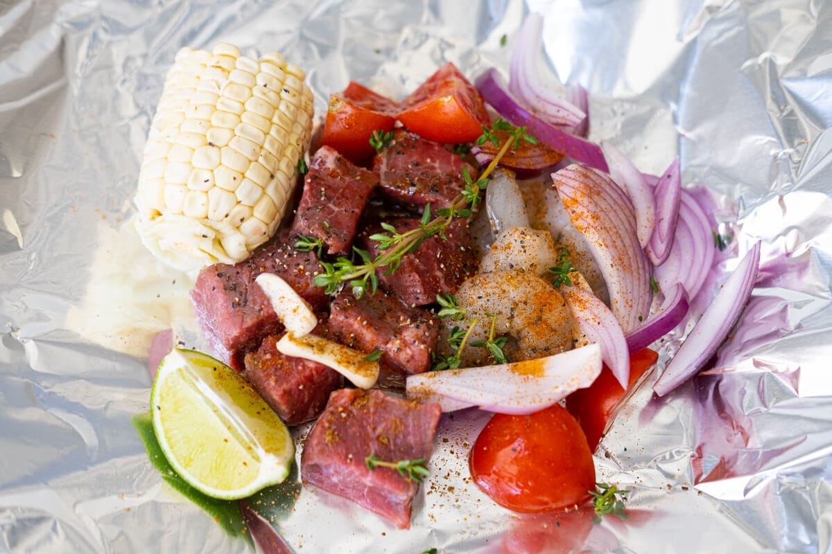 Steak, shrimp and vegetables arranged on foil paper with add seasonings on top.