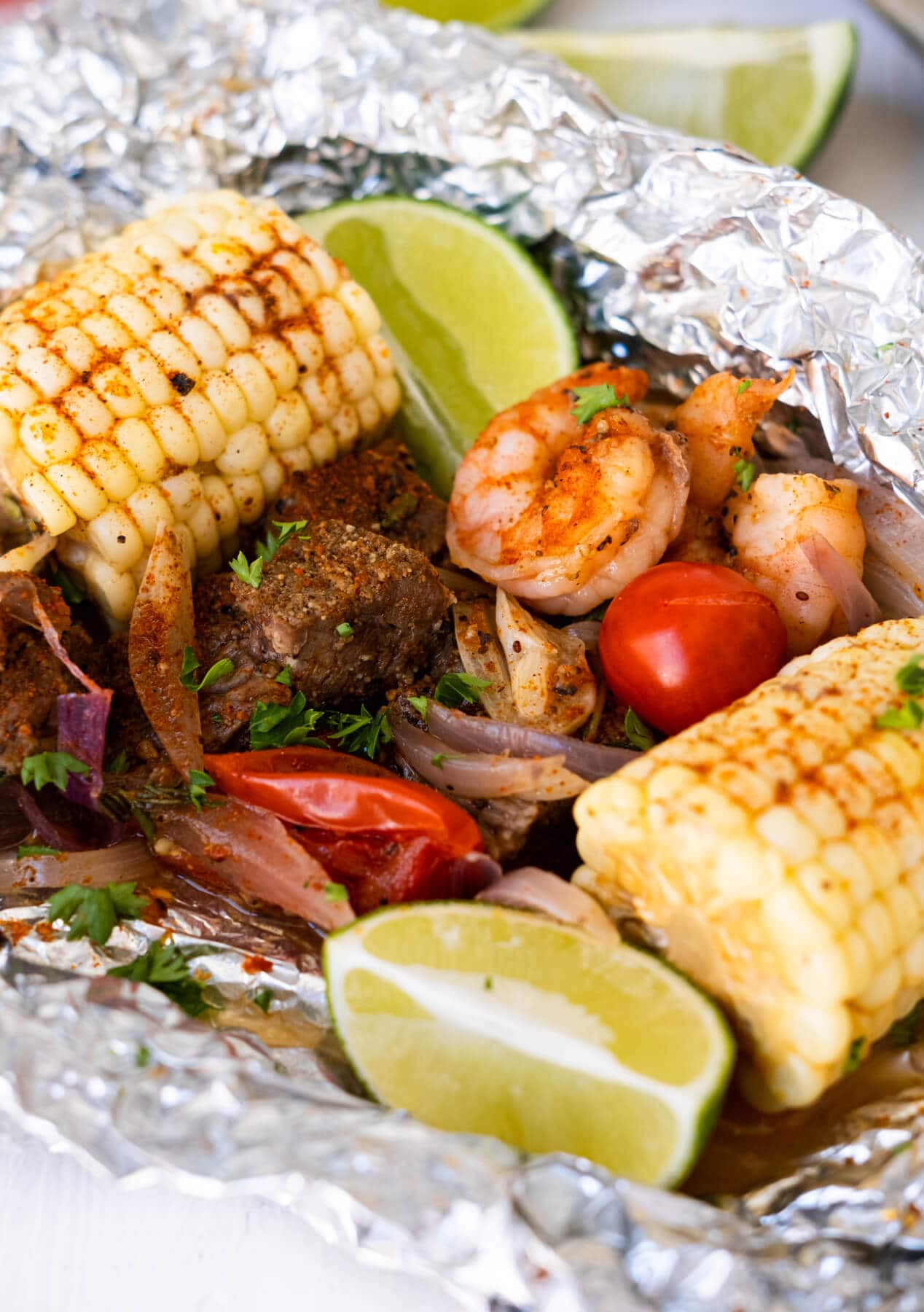 Grilled steak, shrimp, tomatoes, onion slices, corn with lime wedges and topped with chopped parsley on top in tin foil.   