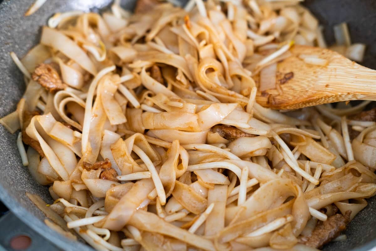 Flat rice noodles being stir fried with the seasoning in the wok with a spatula. 