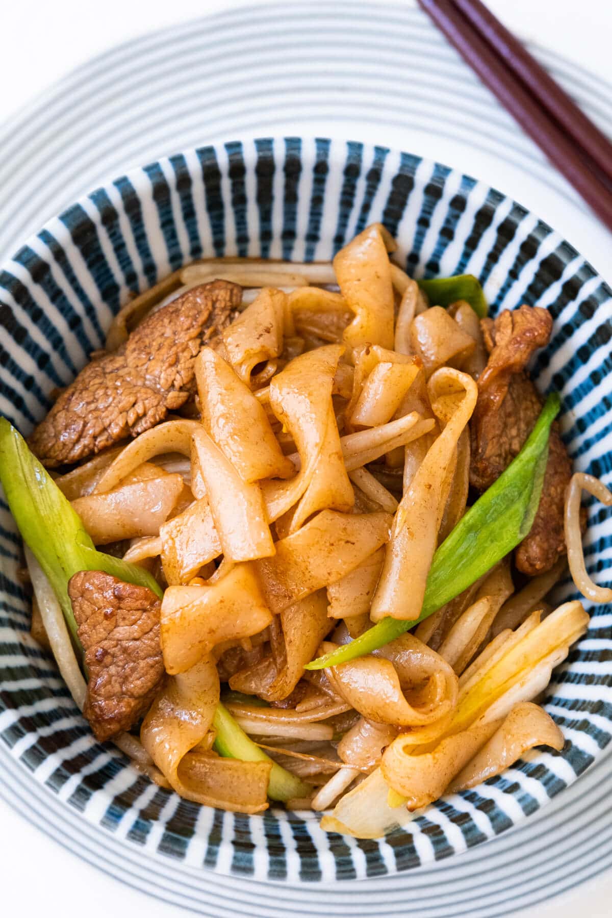 Beef chow fun noodles with beef, green onion and bean sprouts in a bowl with a pair of chopsticks aside. 