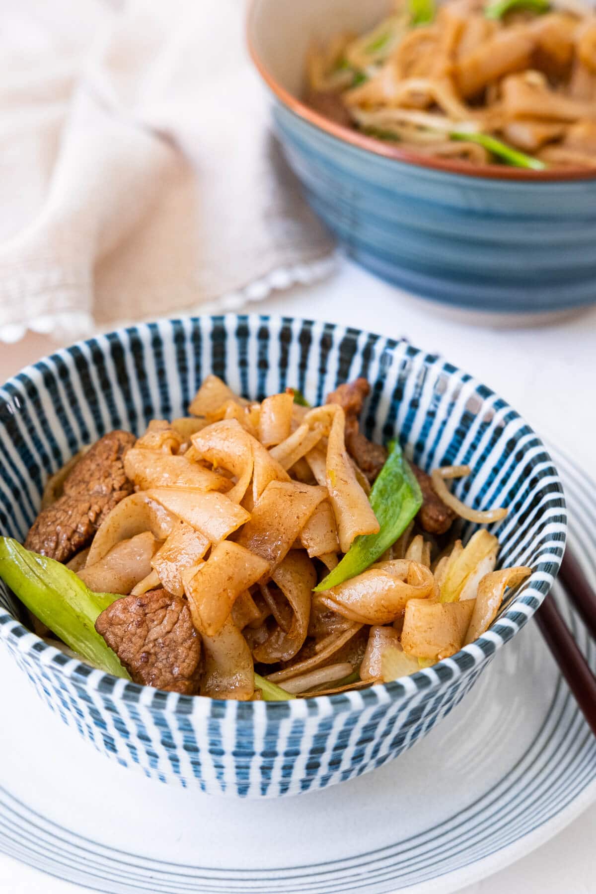 Two bowls of flat rice noodles filled with beef, fresh bean sprouts, and green onions placed on table and with the kitchen towel placed next to them.