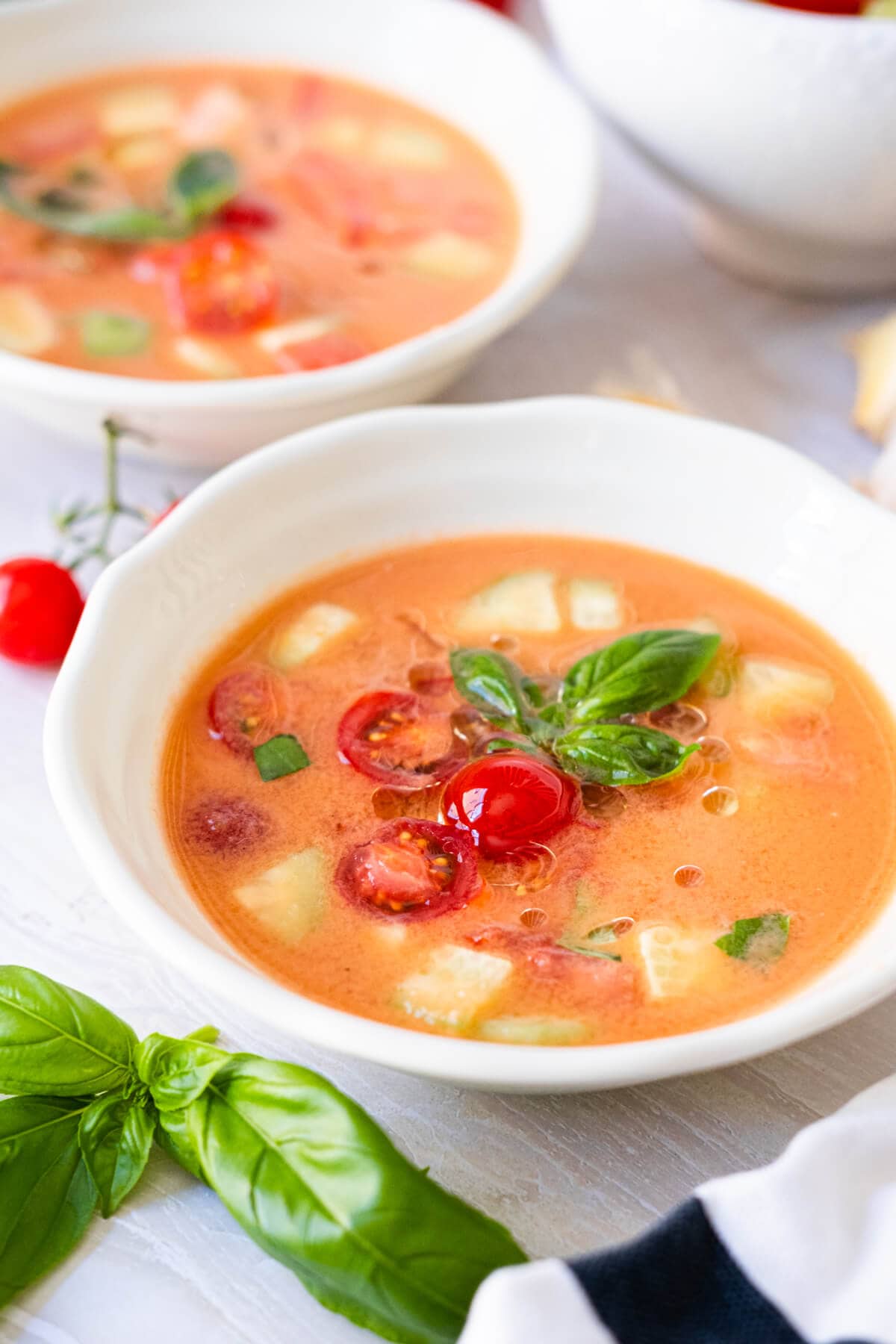 Orange-red  gazpacho served in a white small bowl with tomatoes, cucumber chunks and basil leaves in top. 