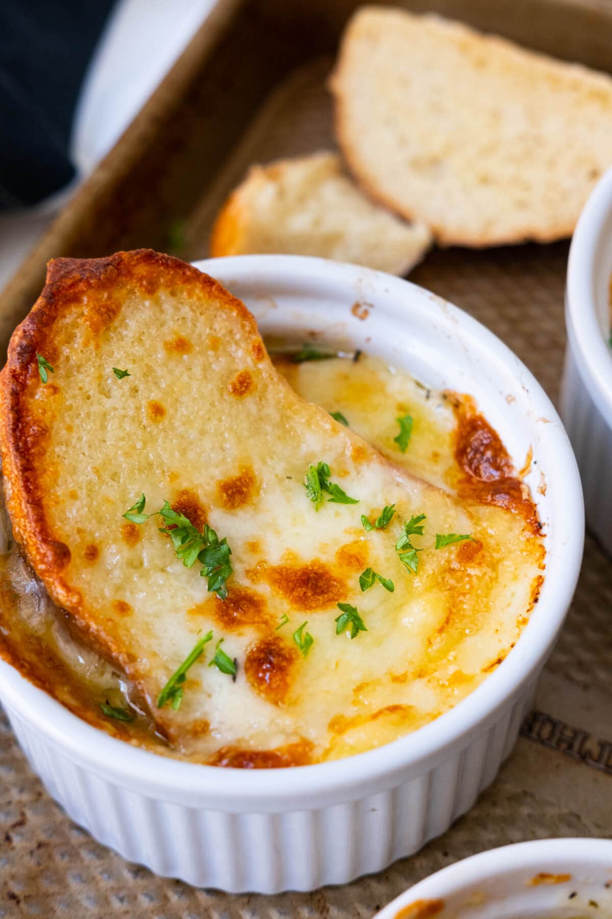 Golden brown broiled soup with a slice of bread dipped within it covered with melted cheese served in a ramekin with two slices of bread aside. 