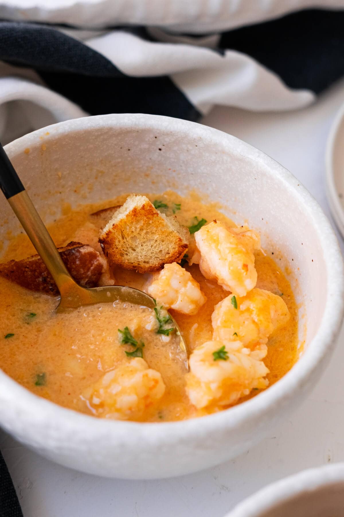 A spoon scooping a spponful of soup from a white bowl filled with shrimps and croutons. 