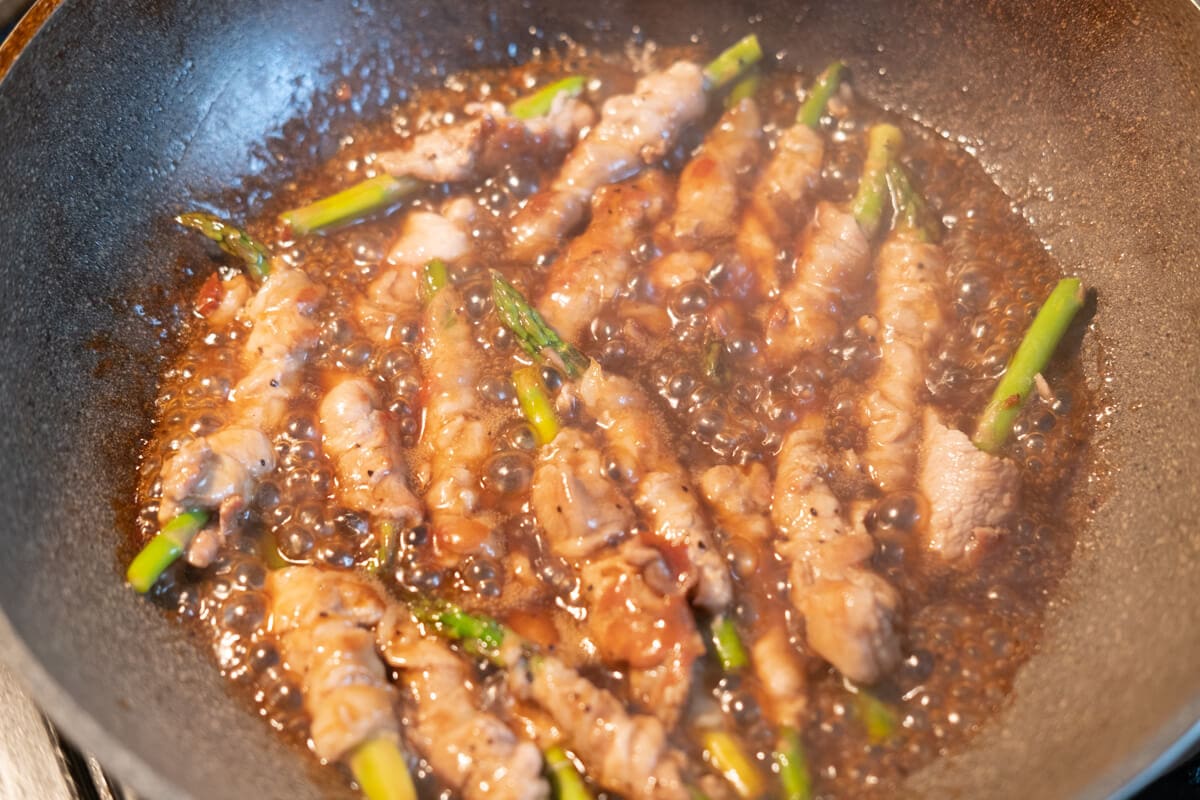 Single pieces of asparagus wrapped around with pork slices simmered in black pepper sauce in a pot. 