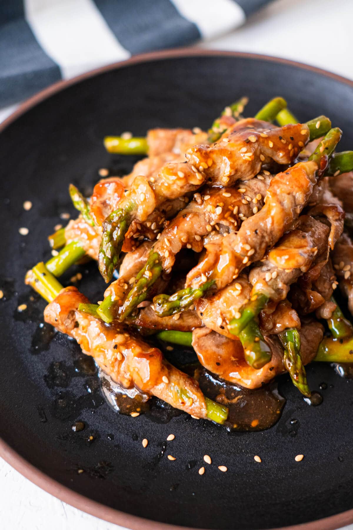 Asparagus with pork wrapped around coated with black pepper sauce served on a black plate with kitchen towel in the background. 