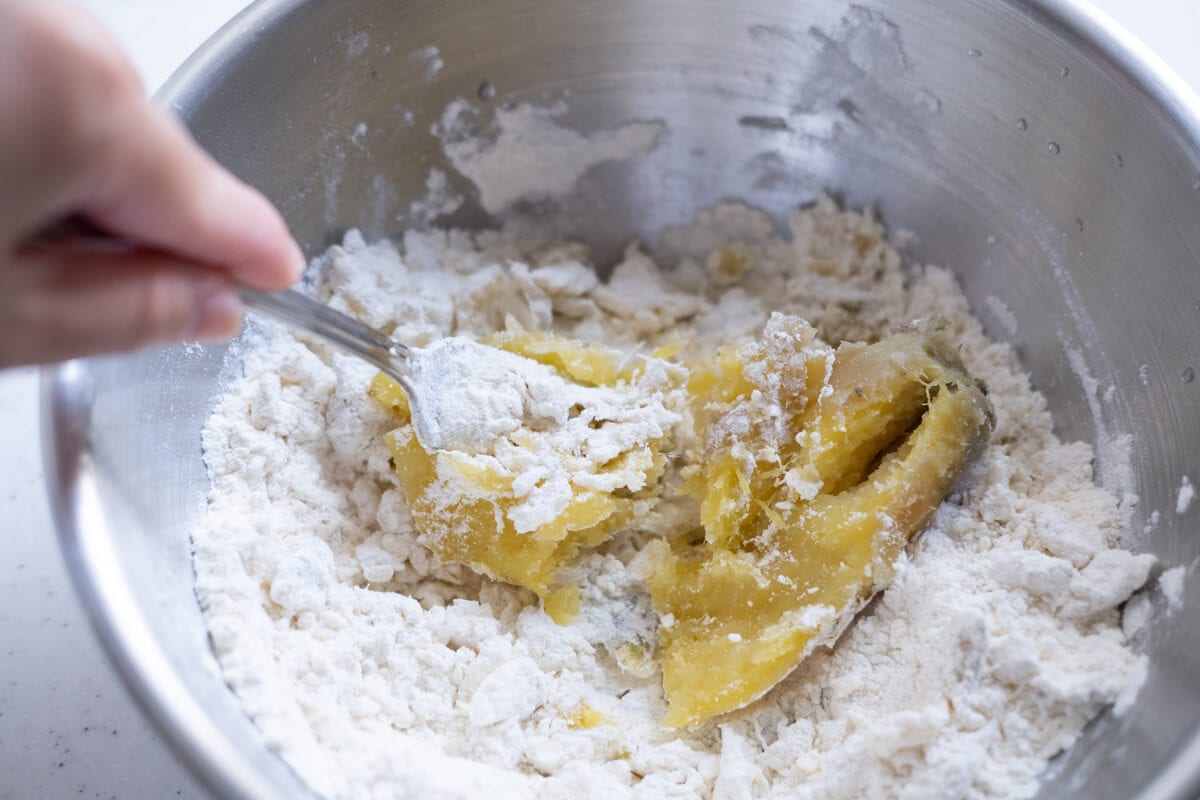 Mashed sweet potato being mixed with all-purpose flour with a fork in a mixing bowl. 