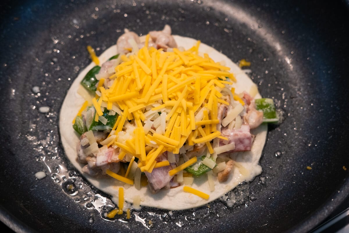 Chicken, bacon, green pepper, and cheese on top of tortilla in a pan. 