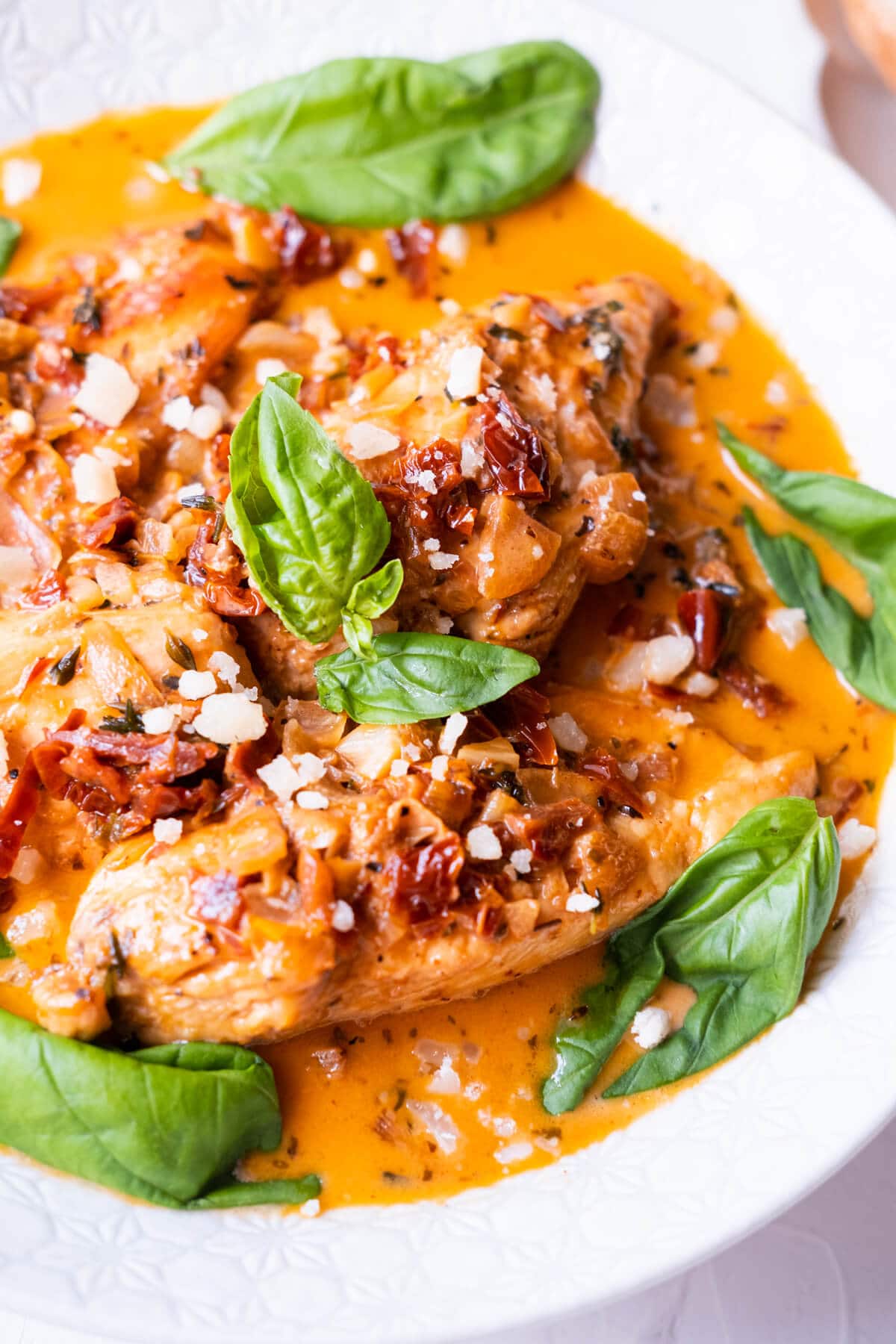 Close up of seared chicken breast slices in a red creamy sauce with chopped sun-dried tomatoes, and topped with basil leaves and grated Parmigiano Reggiano.