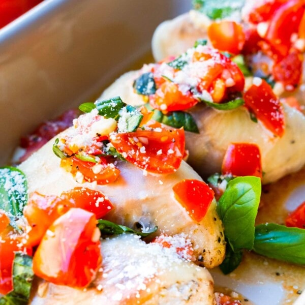 Chicken breast covered with melting cheese and served in a rectangular dish with chopped tomatoes and basil leaves.