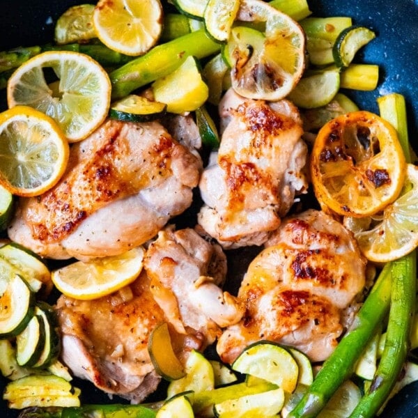 One pan greek chicken cooked in a skillet with asparagus, zucchini slices and lemon slices.