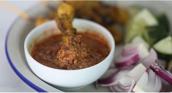 Chicken Satay dipped in a bowl of peanut sauce served on a plate with onions and cucumbers. 