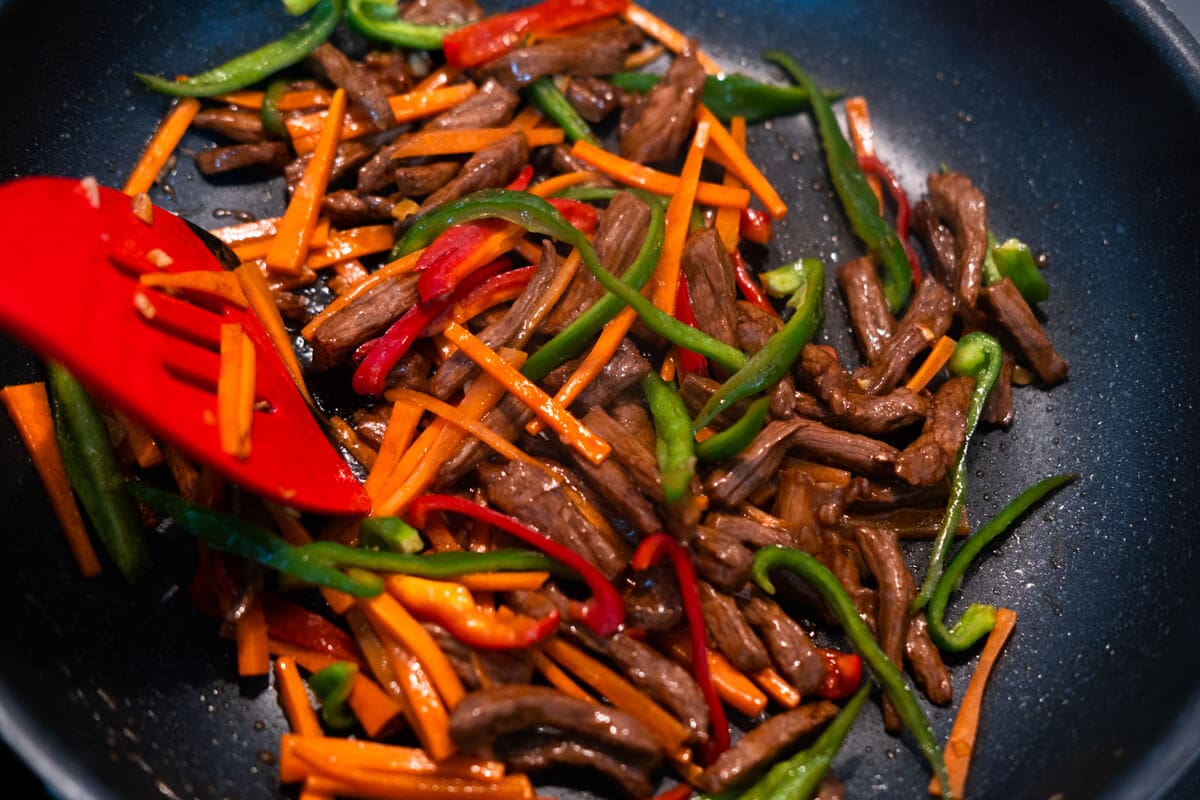 Adding vegetables to beef in a pan. 