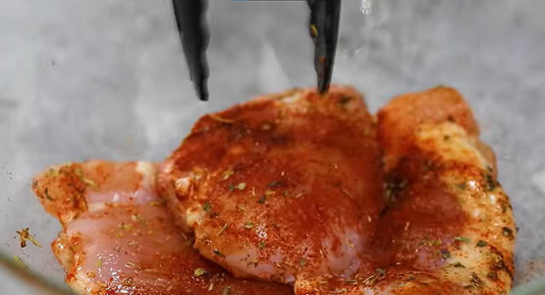 Chicken thighs in a bowl marinated with smoked paprika, Italian seasoning, salt and olive oil mixed using a tong. 