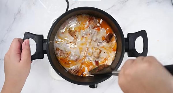 Coconut milk and water added to the skillet with the spice paste, cinnamon, cloves, star anise, cardamom pods, lemongrass and chicken and being stirred with a spatula. 