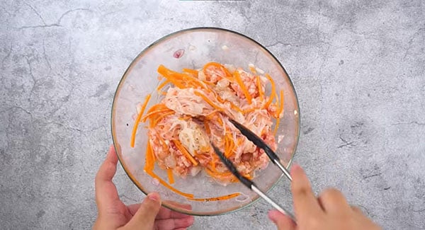 Mung bean noodles, ground pork, shrimp, crab meat, shallot, garlic, egg, carrot, ground black pepper, salt and fish sauce mixed in a large bowl with tongs. 