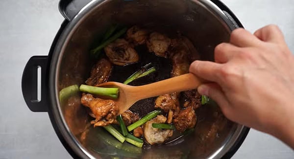 Scallions are being stirred in with the ginger soy chicken using a wooden spoon. 